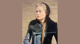 Portrait of filmmaker Flaminia Romani in a black shearling-collar leather jacket with a long braid, photographed against a sunlit brick wall.